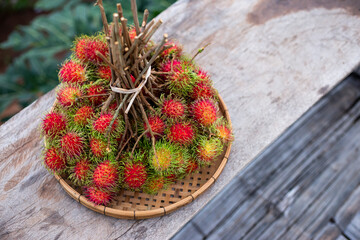 A large bunch of red rambutan in a bamboo tray is placed on a wooden table. Looks delicious to eat rambutan, fresh fruit from the tree in Thailand. Leave space for text