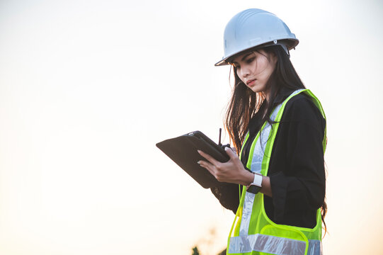 A female engineer is working on a job site and is using a tablet and carrying a walkie-talkie, She is wearing protective workwear and white helmet.