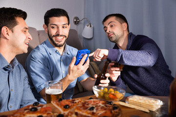 Three happy smiling men using phone while enjoying beer and pizza at home