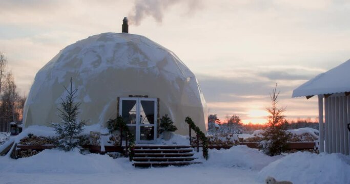 Igloo architecture with steam from chimney at winter evening. Beautiful rural landscape with snow-covered buildings and sunset sky