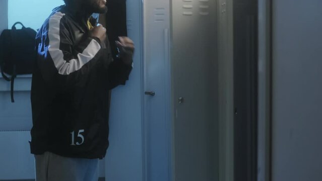 Tilting up of young Black male athlete coming to lockers in dressing room, undressing early in morning
