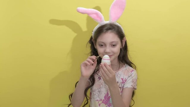 4k. A little girl in rabbit ears paints eggs for Easter on a yellow background in the studio. Hello spring and Childhood lifestyle Happy Easter concept. Easter activities for children