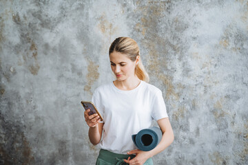 A blonde girl stands with her phone and a yoga mat in her hands. Abstract wall background. Sit on...