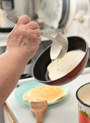 Senior woman chef pour portion of liquid dough by hands with ladle on frying pan for baking delicious pancakes, crepes in the kitchen in the morning for tasty breakfast. Selective focus. Maslenitsa. 