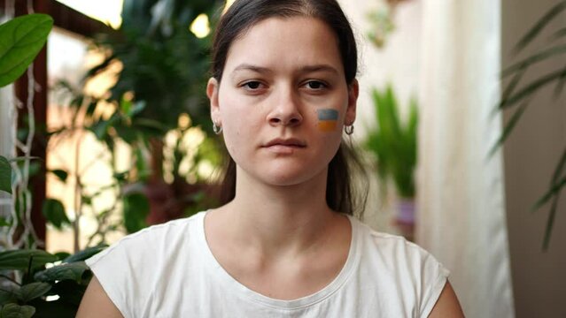 A sad young Ukrainian girl with the flag of Ukraine on her face is looking through window. The concept of participation of the Ukrainian people in the war with Russia. Not war concept