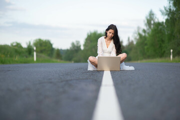 young serious woman sitting on line on asphalt country empty road and using working a laptop computer outdoors