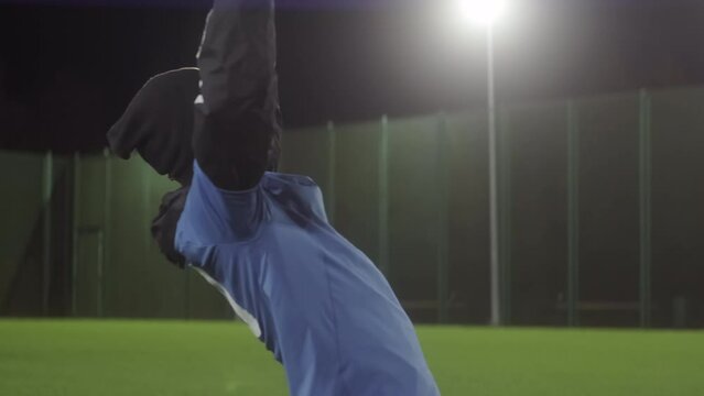 Locked-down of young Black man wearing sports clothing and warm hat, falling on knees on grass on soccer pitch, celebrating goal, imagining being famous player