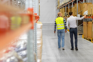 wholesale, logistic business and people concept - manual worker with clipboard and businessman walking along warehouse
