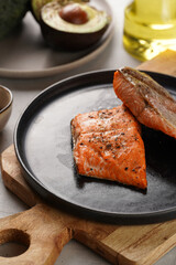 Two grilled fish steaks - red salmon on black round plate on wooden board, olive oil, avocado on grey background