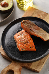 Two grilled fish steaks - red salmon on black round plate on wooden board, olive oil, avocado on grey background