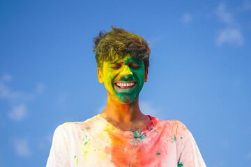 Portrait of a young indian boy playing holi. Face covered in Holi colors with a beautiful blue sky...