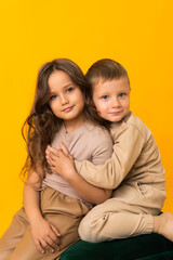 A sister and a brother hugging and looking at the camera. Studio portrait on a yellow background