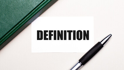 On a light gray background lies a pen, a green notebook and a white card with the text DEFINITION. Business concept