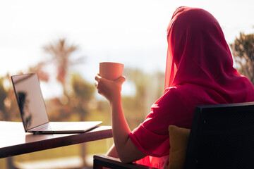 Silhouette of muslim, arab woman in red head scarf, hijab sitting at the table on terrace with...