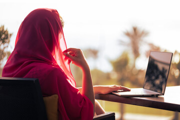 Silhouette of muslim, arab woman in red head scarf, hijab sitting at the table on terrace with laptop opposite sunset palm sea beach of United Arab Emirates