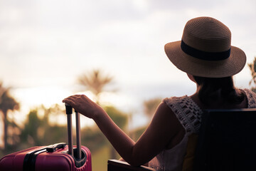 Silhouette of tourist woman in straw hat sitting on the armchair with suitcase on terrace and looking on sunset palm sea beach. Travel,vacation concept.Rear view