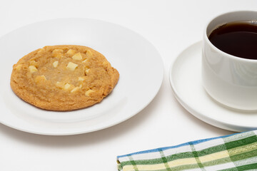 A home-made white chocolate chips cookie on a white plate and a fresh cup of fresh coffee on a white background at a coffee break