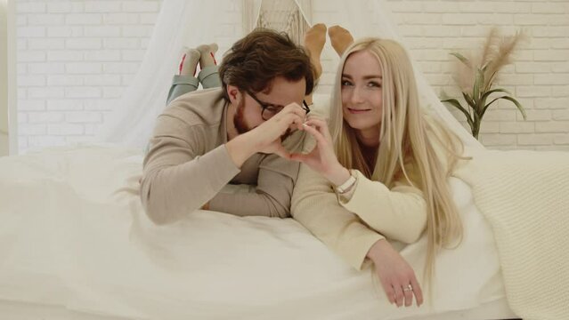 Cute Caucasian Couple Looking At Each Other Forming Heart Shape With Their Palms While Laying On Their Bed. High quality 4k footage