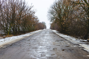 Old broken asphalt road with pits and puddles through winter forest