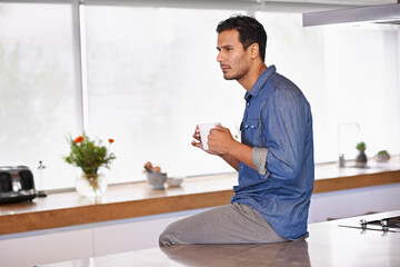 Contemplation over coffee. A serious man holding a mug of coffee and sitting on his kitchen counter.