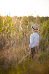 Naklejka na ściany i meble A child in a straw hat on a summer meadow in the rays of the setting sun.