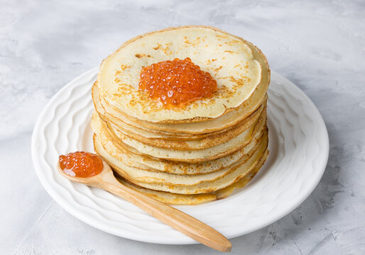 Pancakes for Maslenitsa with red caviar on a white plate and a wooden spoon.