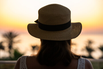 Silhouette of woman in straw hat standing on balcony and looking at sunset palm sea beach. Rear view