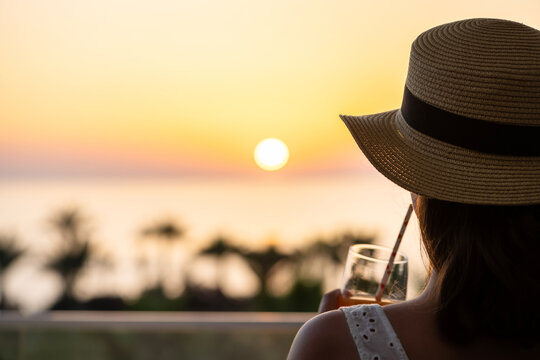 Silhouette of woman in straw hat with juice standing on balcony and looking on sunset palm sea beach. Rear view