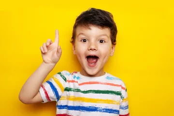 Deurstickers idea. child, boy emotionally points up. A gesture of an idea. Portrait of a child in a bright striped T-shirt on a yellow background © tumskaia