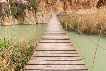 Tibetan bridge made of wood and steel cables that crosses a river towards the wall of the mountain. The green river with a multitude of reeds that grow on the shore. Horizontal photography. 