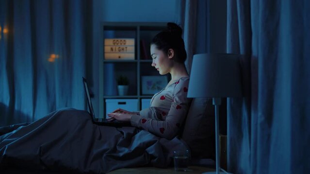 technology, bedtime and people concept - teenage girl with laptop computer sitting in bed at home at night