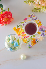multicolored meringues in a glass vase on the table, cup of tea, spring flowers white and pink, top view, copy space