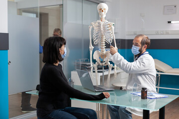 Orthopedic specialist explaining bones injury on human skeleton to woman in medical cabinet....