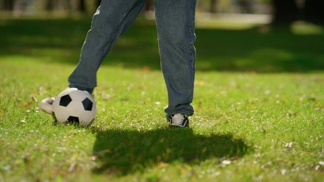 Male legs kicking ball on green field sunny park closeup. Summer active weekend.