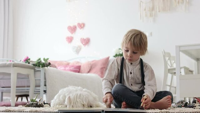 Cute blond preschool child, blond boy with pet maltese dog, reading book at home on the floor with mother