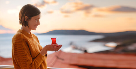 Woman drinking turkish tea from traditional turkish teacup and enjoys panorama over sunset of Kas...