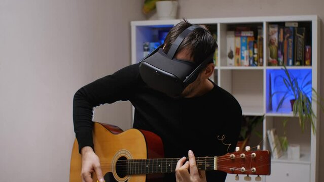 A man wearing black coffee and virtual reality glasses plays an acoustic guitar. Behind him is a rack of games and flowers. VR helmet for learning music and online concerts