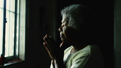 Spiritual older woman in 80s praying at home by window