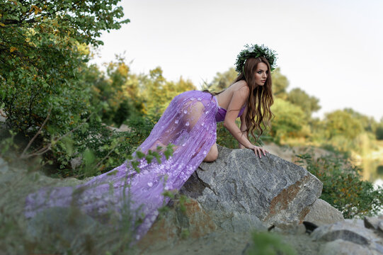 Mavka water nymph naiad near the water in nature in the forest on the river bank of the lake on a quarry on a cliff in a purple lilac transparent dress with a wreath on her head