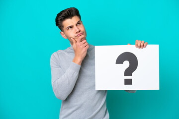 Young handsome caucasian man isolated on blue bakcground holding a placard with question mark symbol and thinking