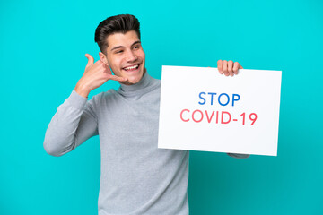 Young handsome caucasian man isolated on blue bakcground holding a placard with text Stop Covid 19 and doing phone gesture