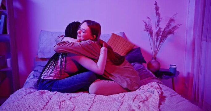 Happy meeting of two friends hugging in the room sitting on the bed. Smiling girls friends laughing and hugging. Multiethnic young women embrace each others after a long time they have been distant.
