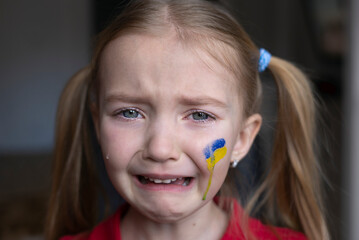 the face of a frightened girl, a child crying, tears flowing, painted on her cheek in the yellow-blue colors of the Ukrainian flag, a request for help. Children ask for peace