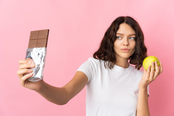 Teenager Ukrainian girl isolated on pink background having doubts while taking a chocolate tablet...