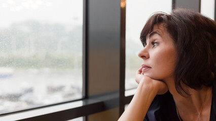 Woman 35-40 years old sadly looks at rain outside window,