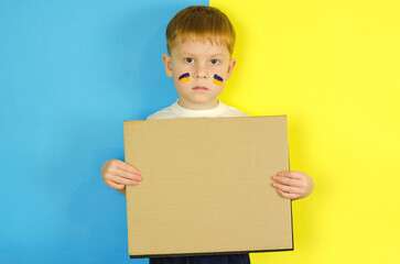 A Ukrainian boy stands against the background of the Ukrainian flag and holds an empty poster, space for text. Sad child with painted flags of Ukraine on his cheeks