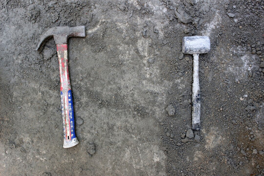 Overhead View Of A Used Standard Claw Hammer And A Plastic Mallet On A Grey Dirt Background. No People.