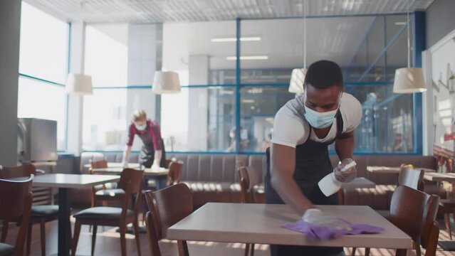 African-American waiter in safety mask and gloves clean table in cafe. Realtime