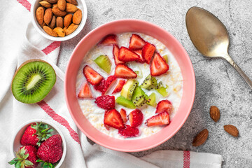 oatmeal porridge with slices of kiwi, strawberries, almonds in pink bowl, spoon, napkin with red stripes on concrete background Vegan food concept Healthy breakfast