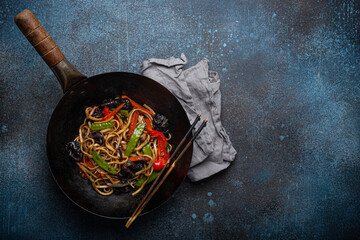 Asian dish stir fry udon noodles with vegetables and mushrooms in black rustic wok pan with wooden chopsticks on rustic dark blue concrete background from above, Chinese fast food, space for text
